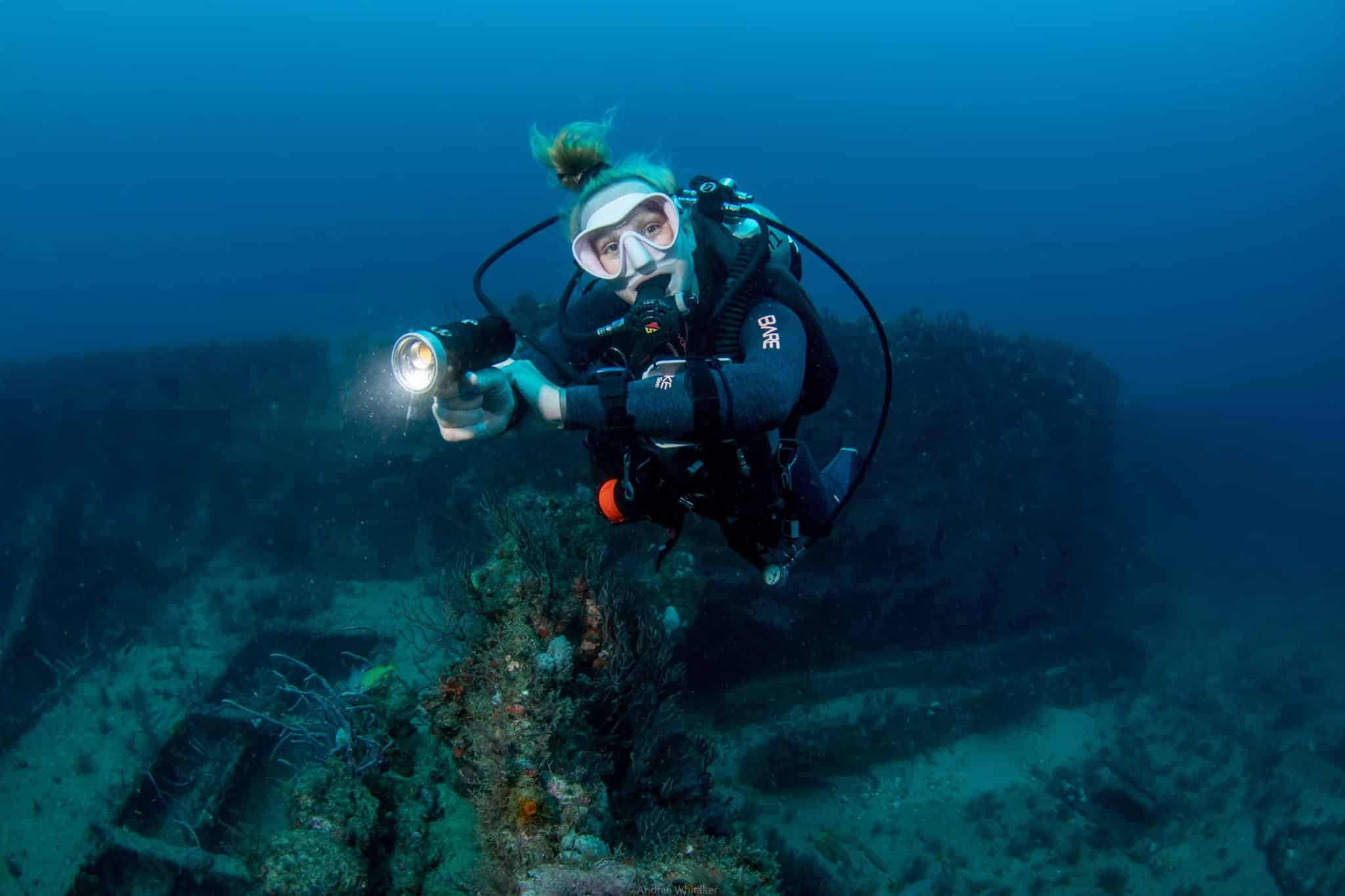 a master scuba diver hovers in front of a wreck as she shines a flashlight