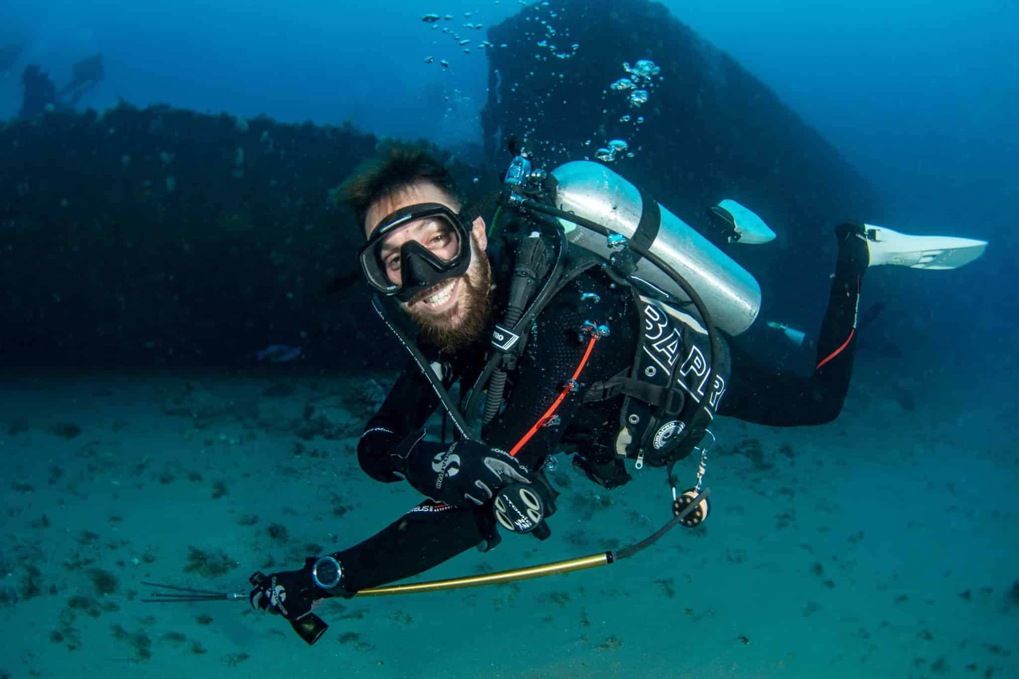 a master scuba diver smiles at the camera as he holds a pole spear in front of a wreck